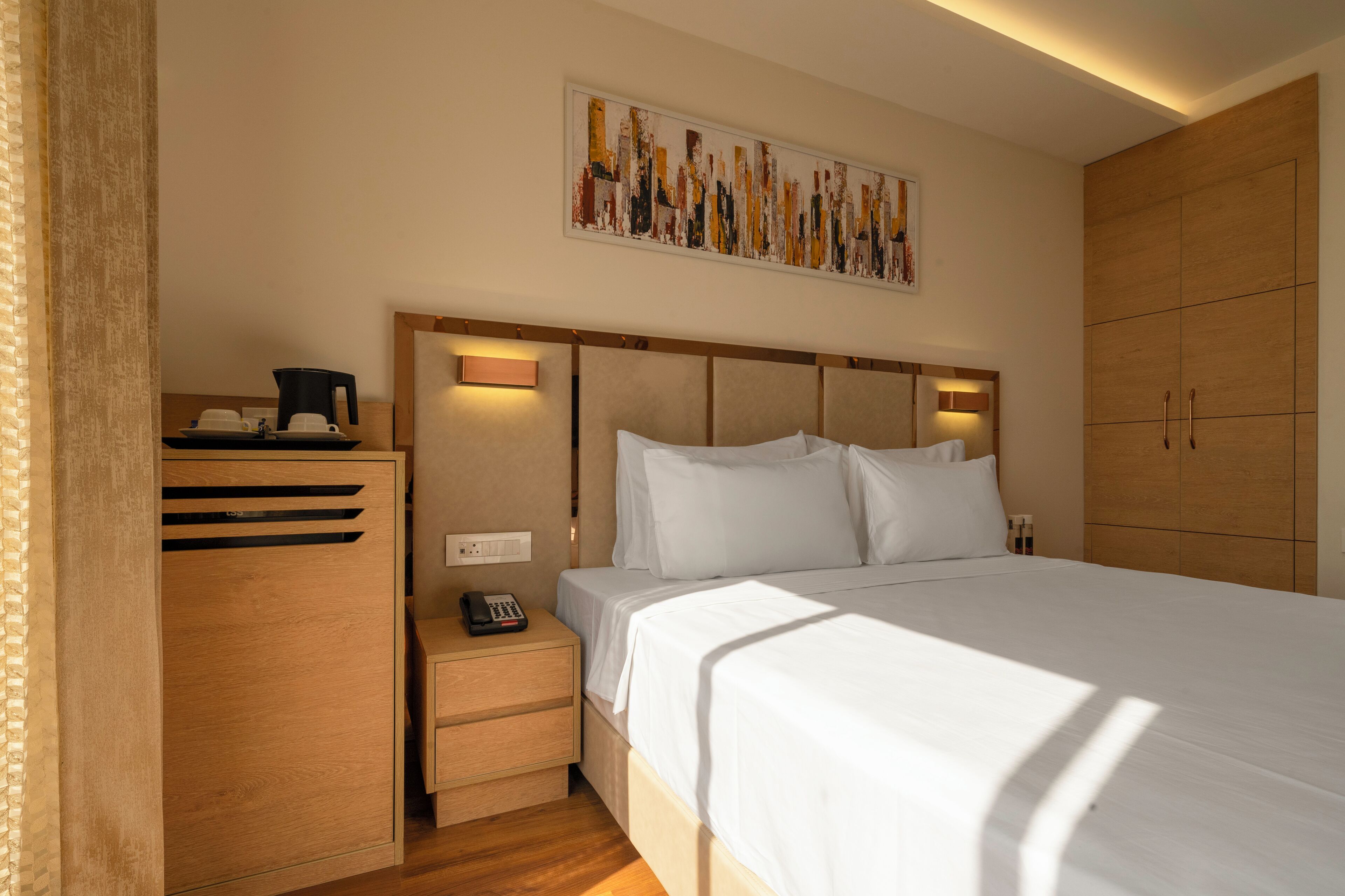 A hotel room with a large bed, a wooden wardrobe, a nightstand with a phone, and a painting on the wall.