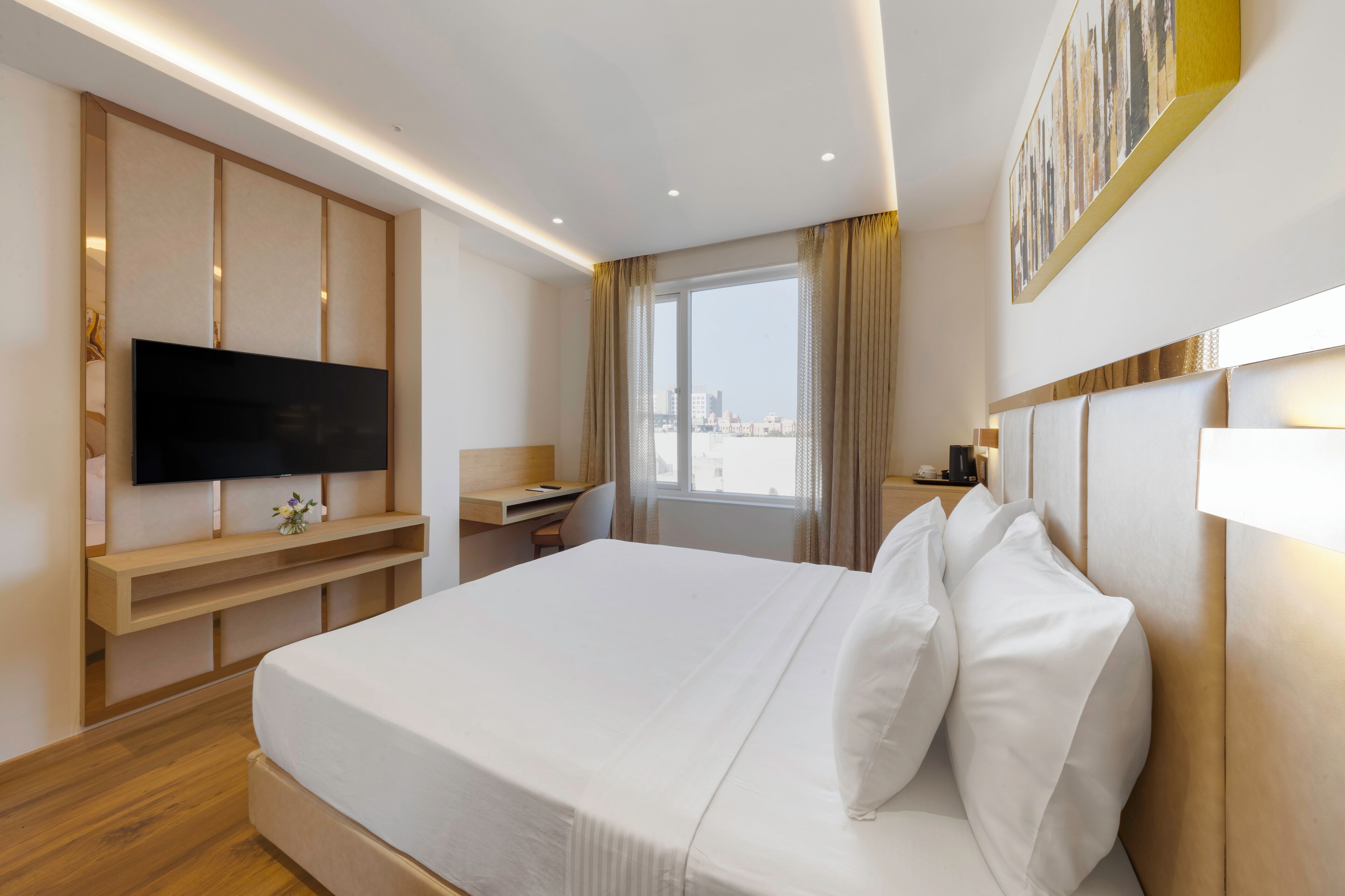 A hotel room with a large bed, a TV mounted on a wooden panel, a desk, and a window with curtains.