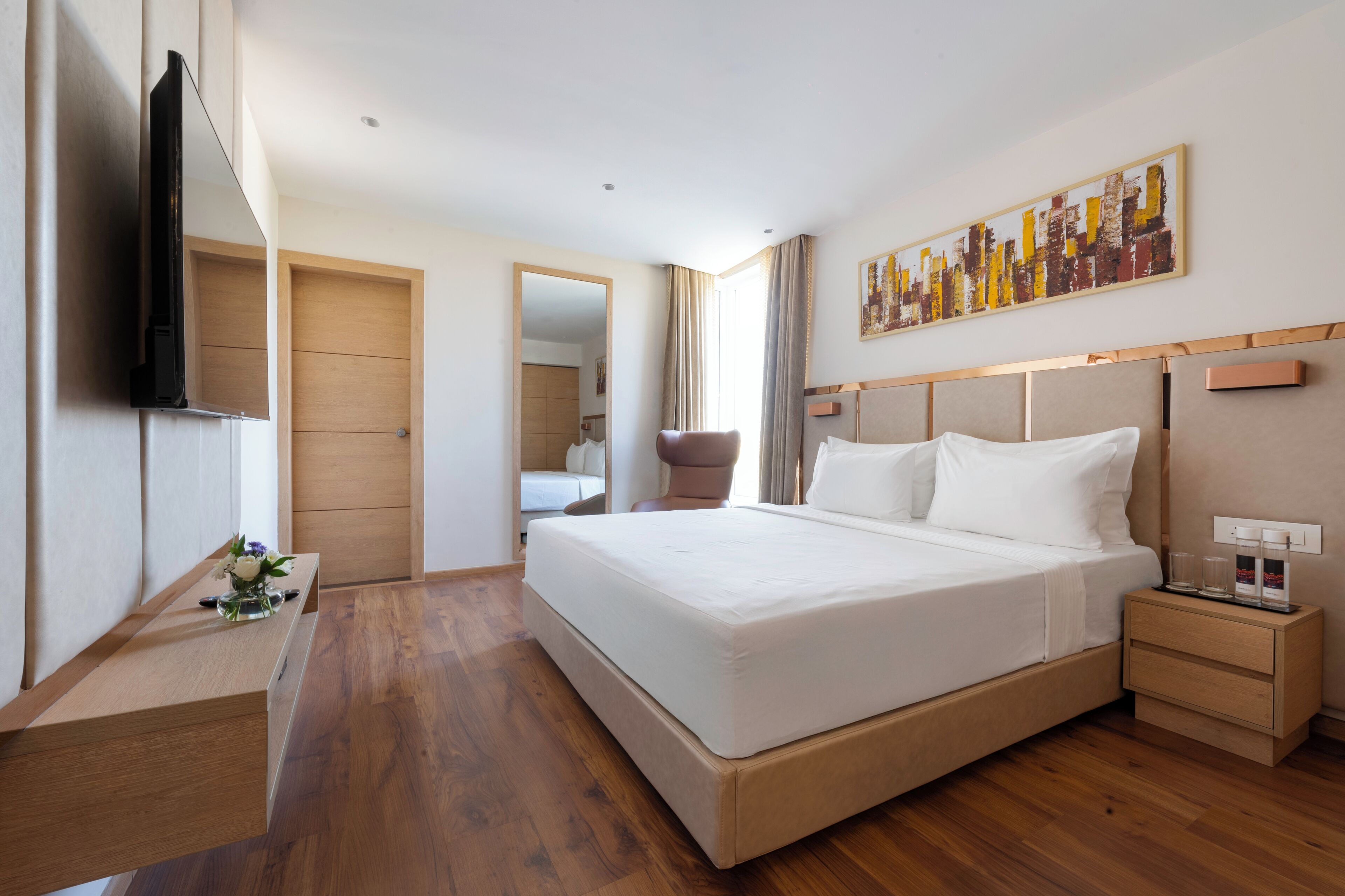 A modern hotel room with a large bed, wooden flooring, a flat-screen TV, a bedside table with glasses, and a cityscape painting on the wall.