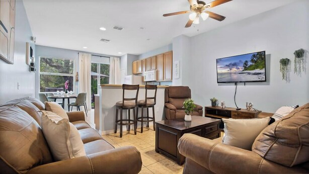A modern living room with leather sofas, a dining area with chairs, a kitchen with wooden cabinets, and a TV mounted on the wall. - Family escape near Disney parks w/ resort access (Kissimmee)