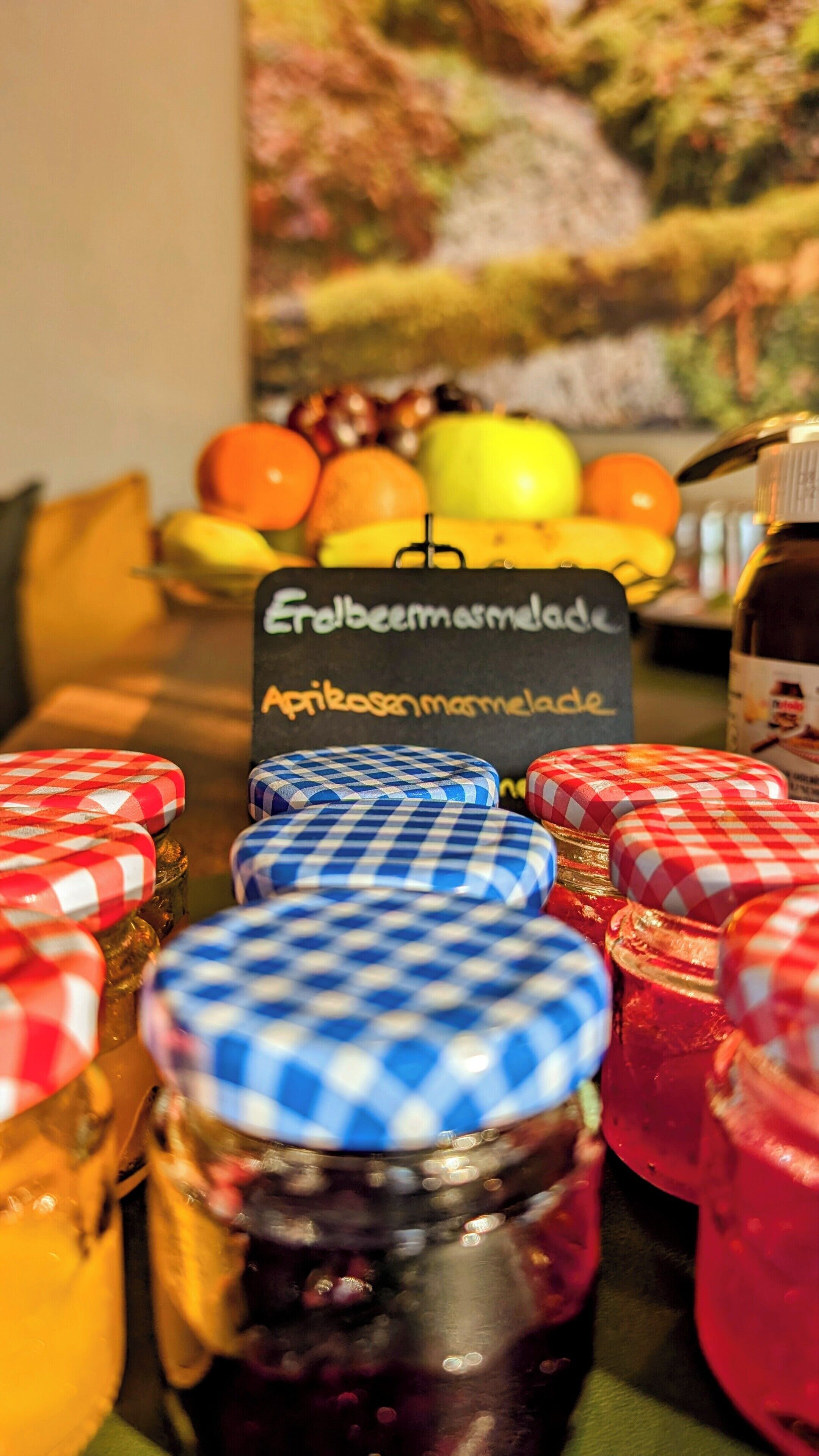 Ein Tisch mit Marmeladengläsern, ein Schild mit Schriftzug und im Hintergrund Obst sowie ein Gemälde.