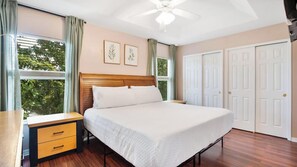 Un dormitorio con cama, mesita de noche, ventilador de techo, ventana con cortinas y puerta.