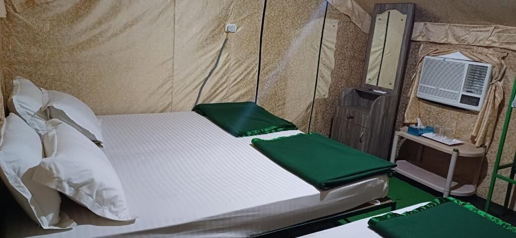 A single-bed room with a green bedspread, white pillows, a bedside table with a mirror, a small cabinet, and a window with an air conditioning unit.