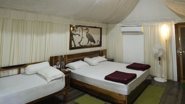 A room with two beds, a wooden headboard, a wall-mounted air conditioning unit, a floor fan, and a painting of a bird on the wall. - SandPebbles Bhitarkanika Jungle Resorts (Rajnagar)