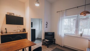 A modern kitchen with a built-in stove, a dining area with a wooden table and black chairs, a TV on a stand, and a large window with curtains.