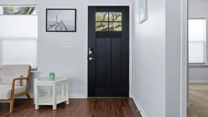 Ein Zimmer mit einer schwarzen Tür, einem gerahmten Bild an der Wand, einem kleinen weißen Beistelltisch und einem Holzstuhl.