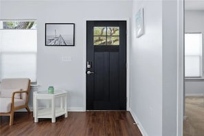 A room with a black door, a framed picture on the wall, a small white side table, and a wooden chair.