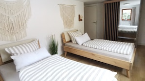 A room with two single beds, a wooden floor, a white wall with macrame decorations, and a sliding door leading to a balcony. - Garden view apartment with pool and children's playground (Dippoldiswalde)
