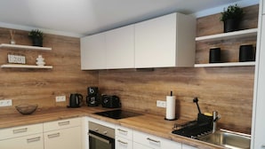 A modern kitchen with white cabinets, a wooden backsplash, a black stove, a sink, and a coffee maker. - Garden view apartment with pool and children's playground (Dippoldiswalde)