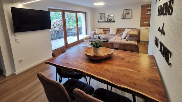 A modern living room with a wooden dining table, comfortable seating, a flat-screen TV, and a balcony with a view of greenery. - Garden view apartment with pool and children's playground (Dippoldiswalde)