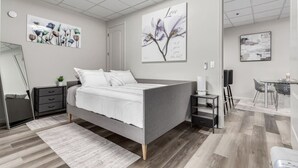 A modern bedroom with a grey bed, a bedside table, a small desk, and a chair. There are two wall-mounted paintings and a mirror. - Stylish & Prestigious 2 Bed 2 Bath-On The Avenue (Fountain Hills)