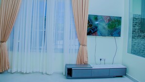 Ein Zimmer mit großem Fenster, leichten weißen Vorhängen und einem beigefarbenen Gardine. An der Wand ist ein Flachbildfernseher angebracht, darunter befindet sich eine kleine Kommode.
