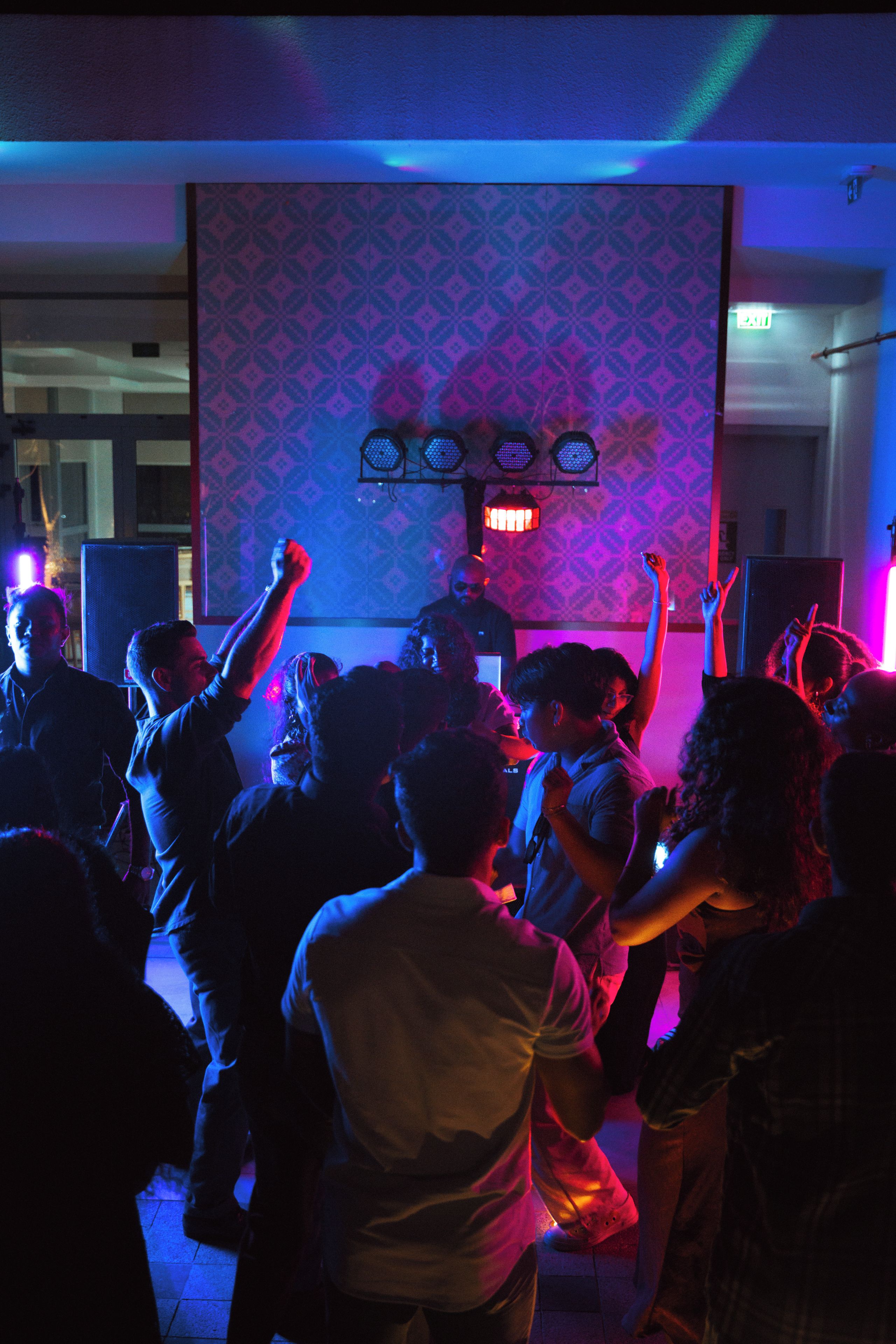 A group of people dancing in a room with patterned wallpaper and a stage with speakers.