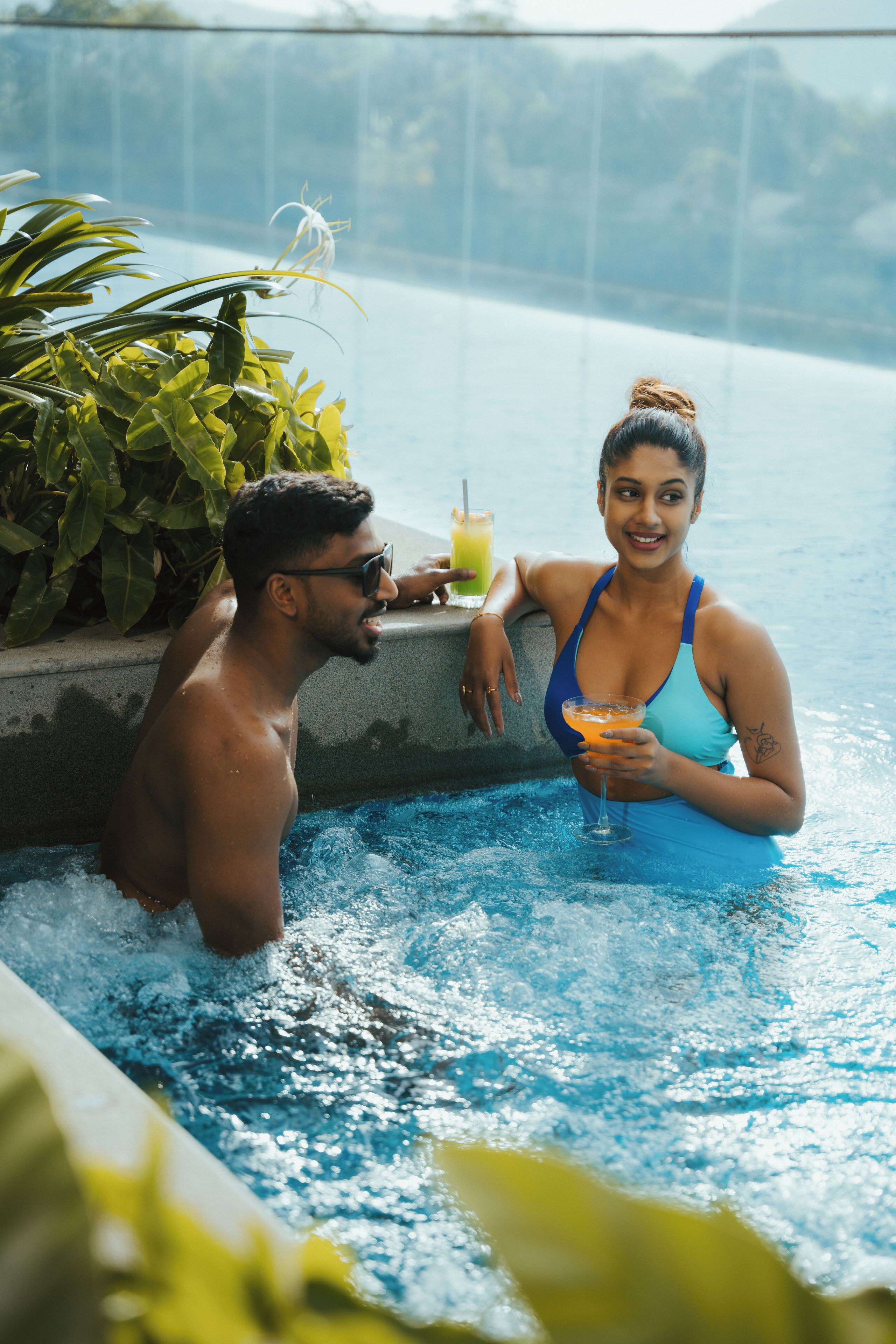 Un couple dans une piscine, avec un verre à la main et une plante tropicale en arrière-plan.