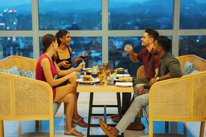 A group of people seated at a table with food and drinks, in a room with large windows and wicker furniture.