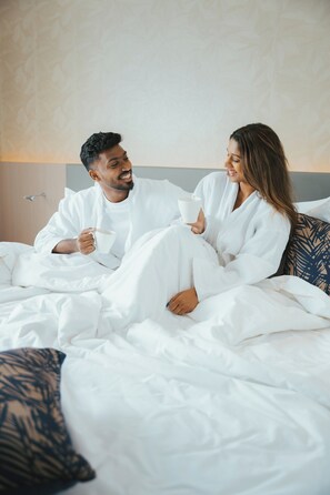 A couple in white robes sitting on a bed with a patterned pillow, holding mugs. - Kandy Myst by Cinnamon (Kandy)