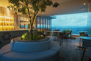 A modern bar area with a seating arrangement, a large tree planter, and a view of the cityscape.