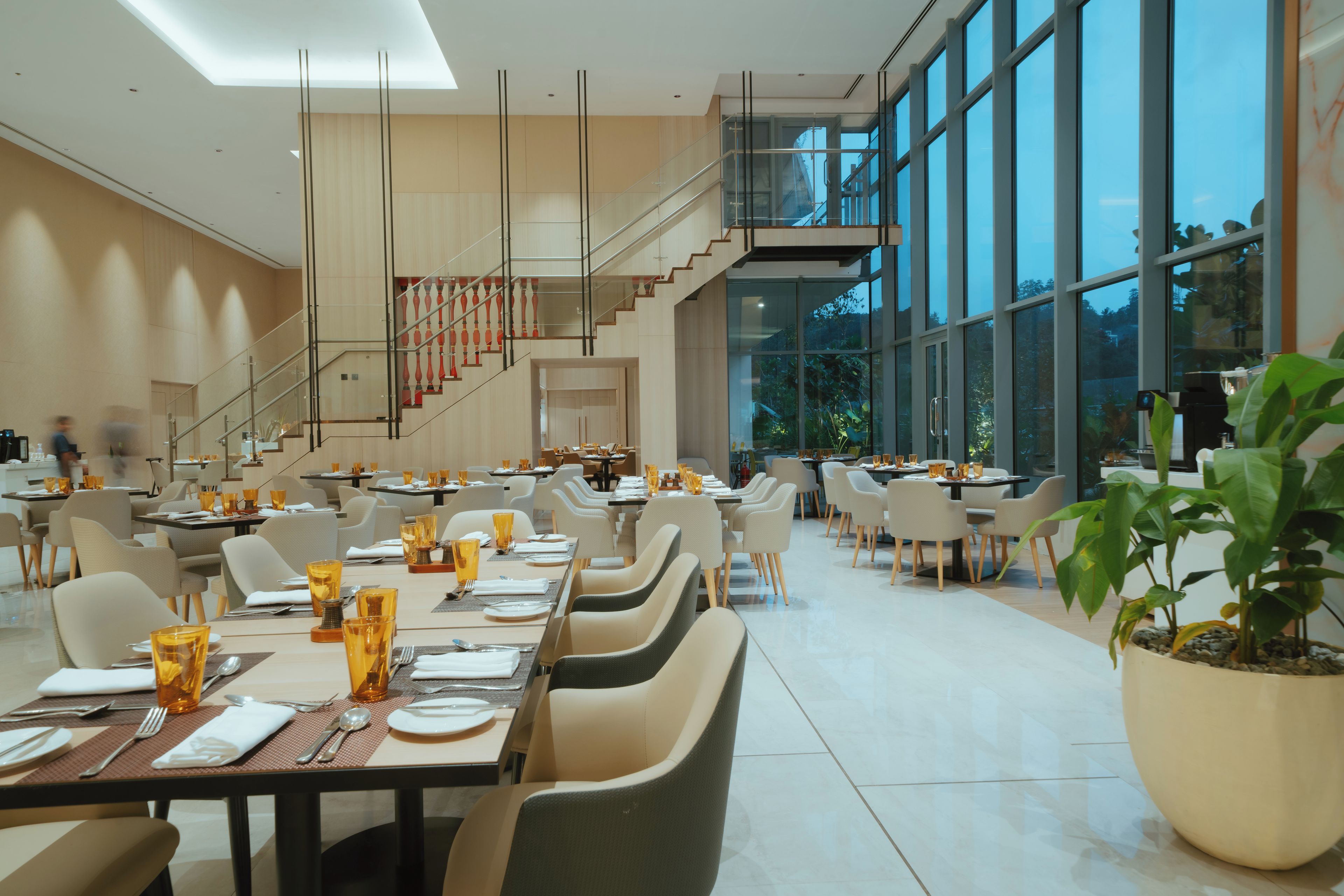 A modern dining area with multiple tables and chairs, a staircase, and large windows offering a view of the outdoors.