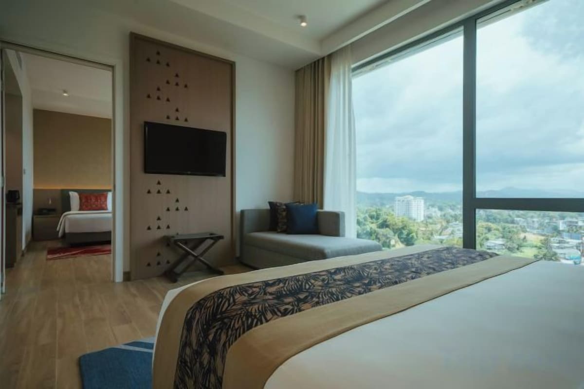 A modern hotel room with a large bed, a TV mounted on the wall, and a view of the city through large windows.
