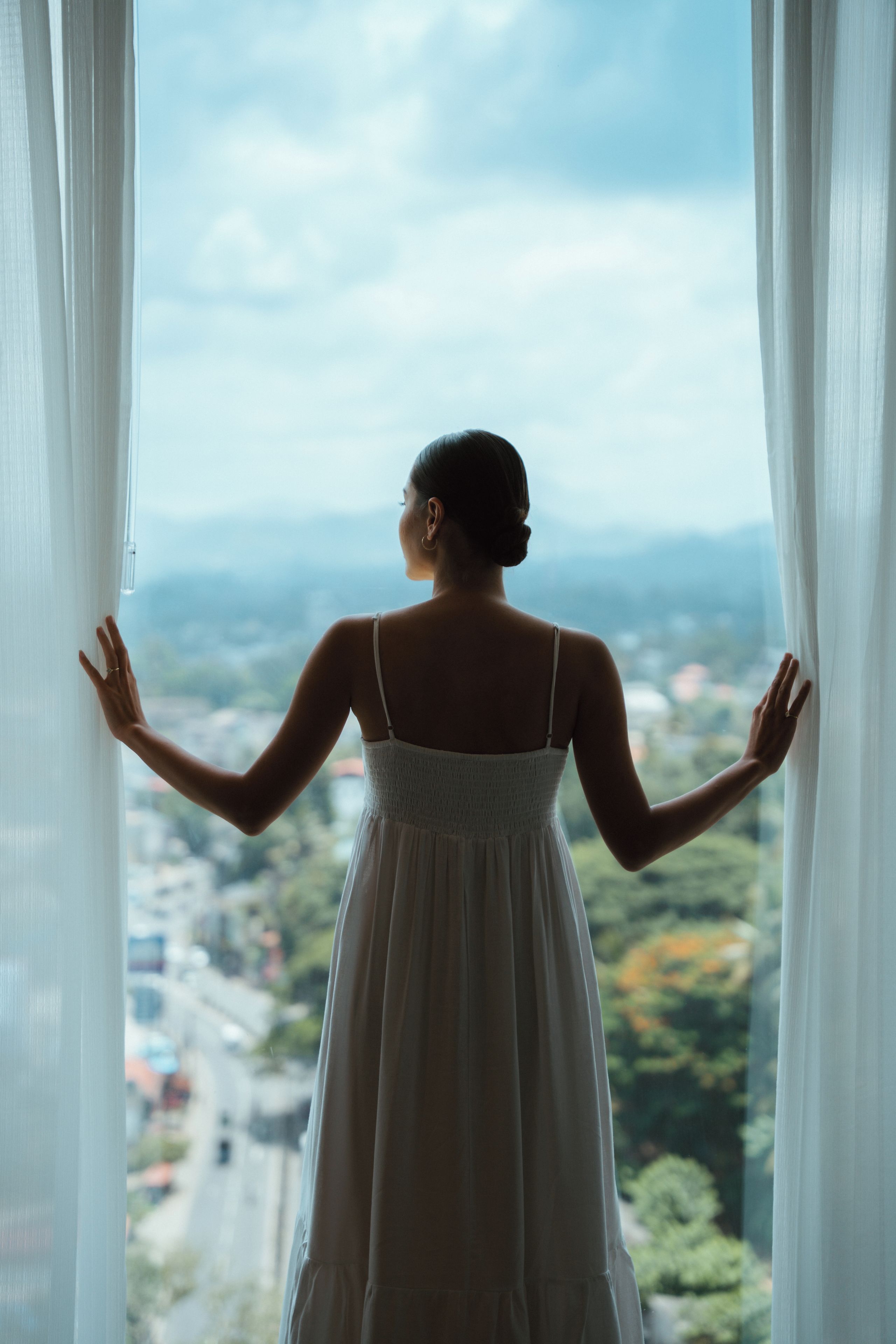 A person standing by a window with curtains, looking out at a view of a cityscape.