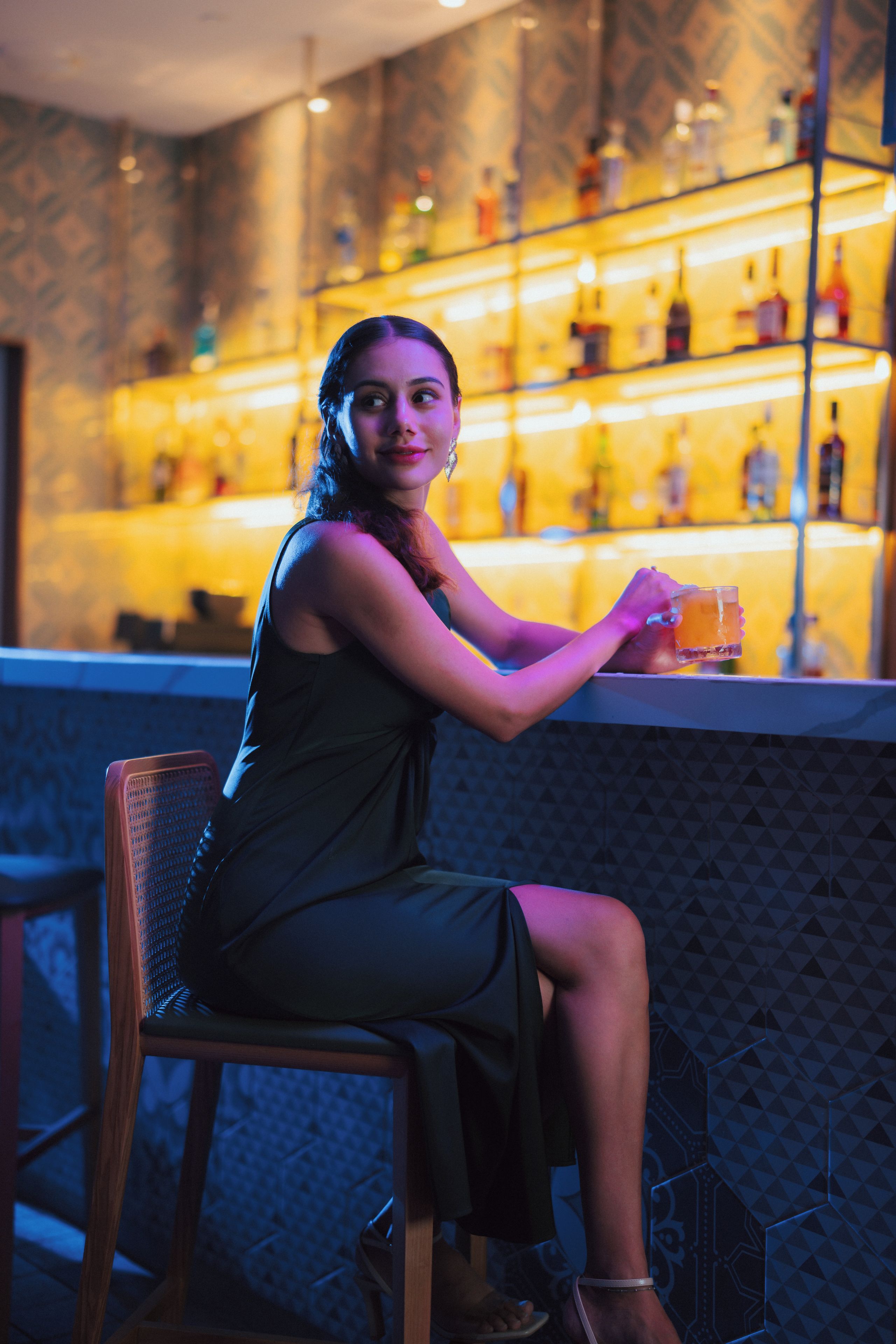 Una persona sentada en la barra con una bebida, con estantes llenos de botellas al fondo.