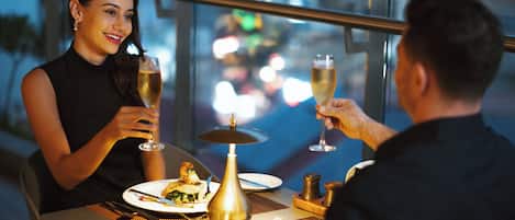 A couple dining at an outdoor table with a city view, holding wine glasses.
