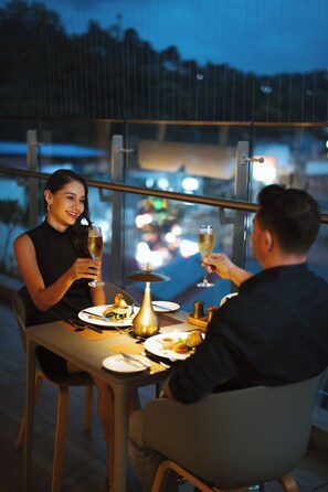 A couple dining at an outdoor table with a city view, holding wine glasses.