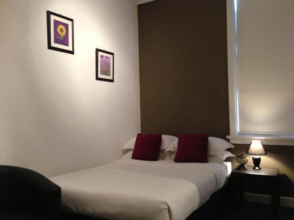 A hotel room with a bed, two pillows, a nightstand with a lamp, and two framed pictures on the wall.