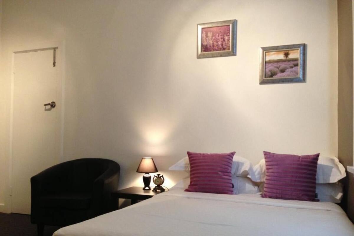 A hotel room with a bed, two pillows, a chair, a nightstand with a lamp, and two framed pictures on the wall.