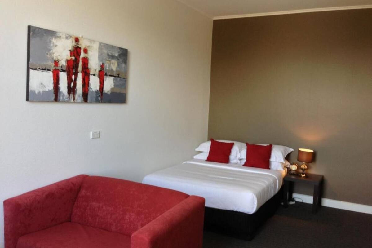 A hotel room with a bed, a red armchair, a nightstand with a lamp, and a painting on the wall.