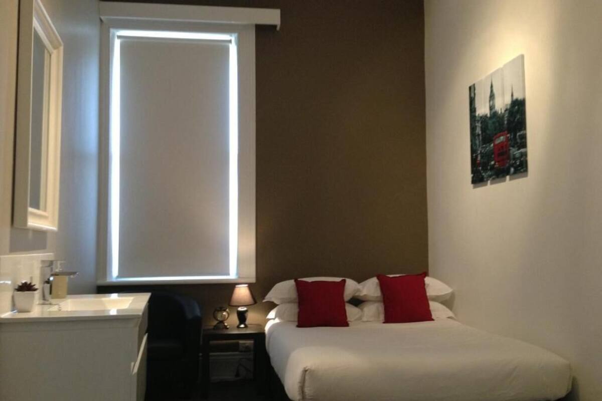 A hotel room with a bed, a nightstand, a lamp, a window with blinds, and a wall-mounted picture.