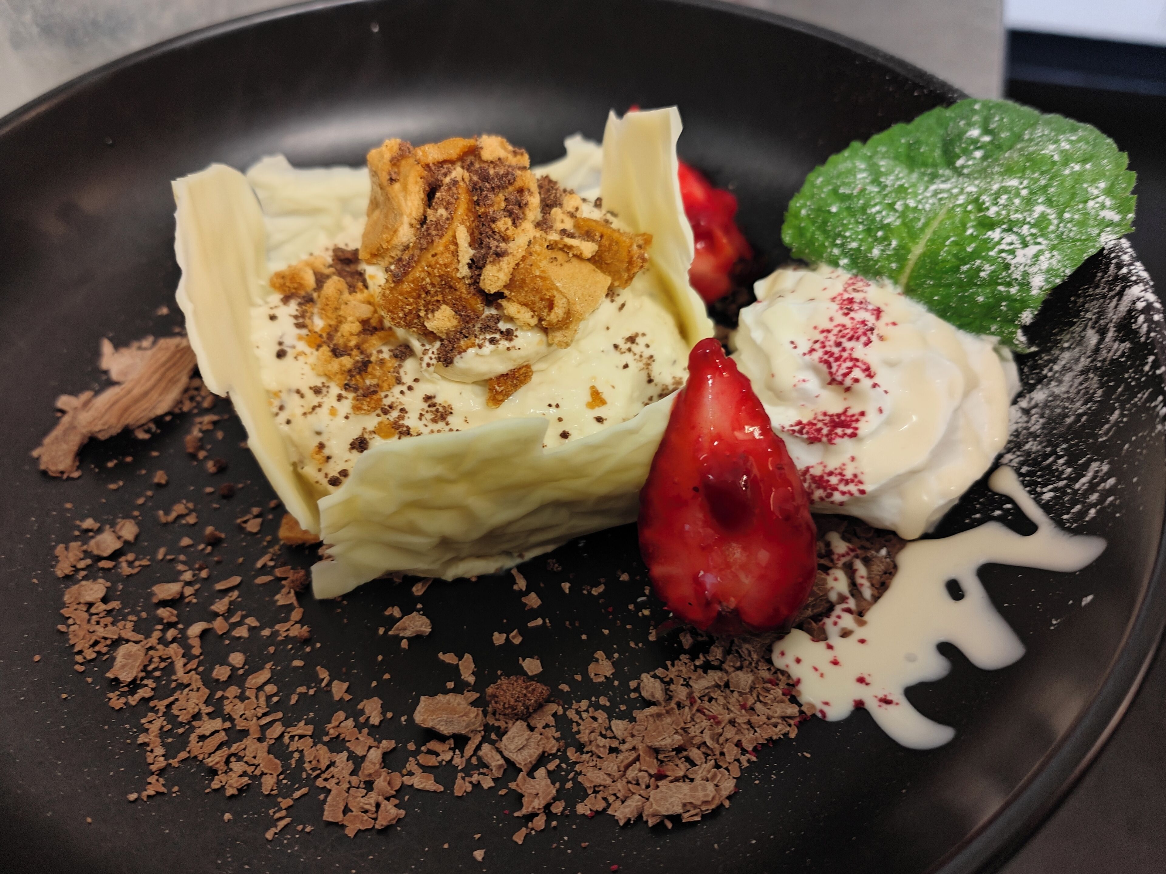 A dessert with whipped cream, crumbled cookies, and a strawberry on a black plate.