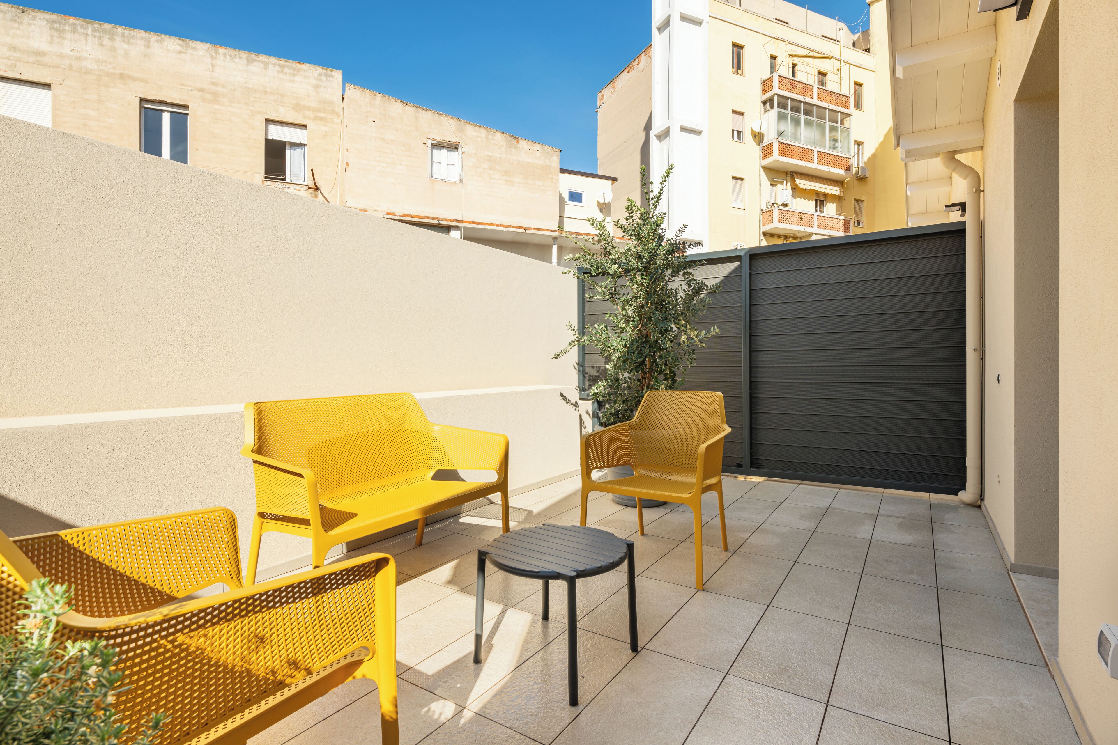 A small outdoor patio with yellow chairs and a table, a closed gate, and buildings in the background.