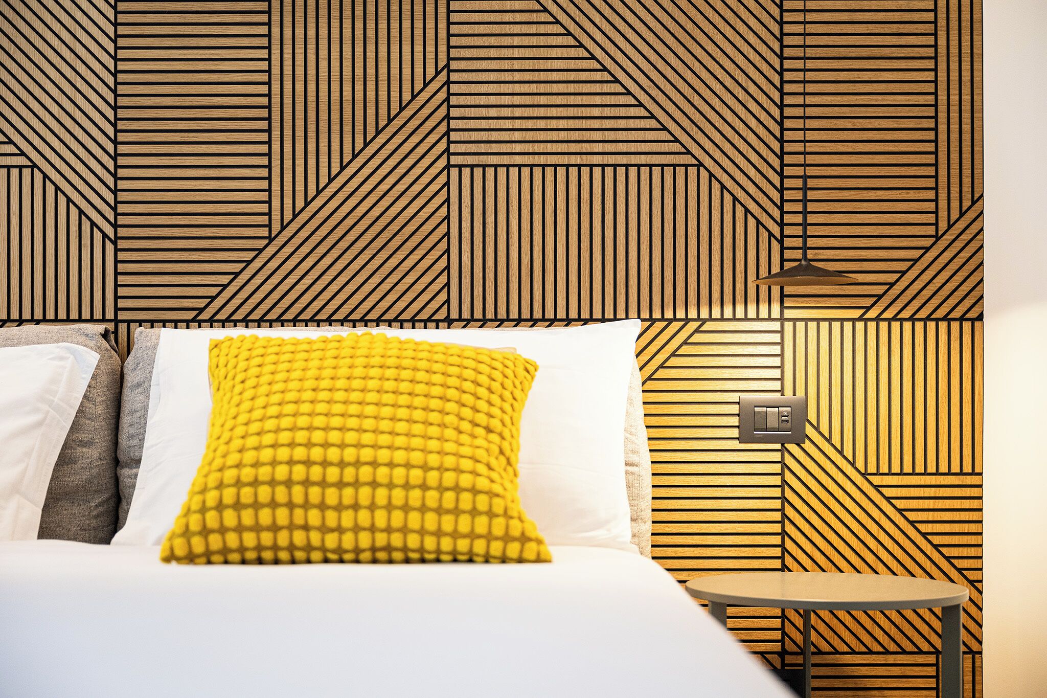 A hotel room with a geometric patterned wall, a bed with white and yellow bedding, and a small round table.