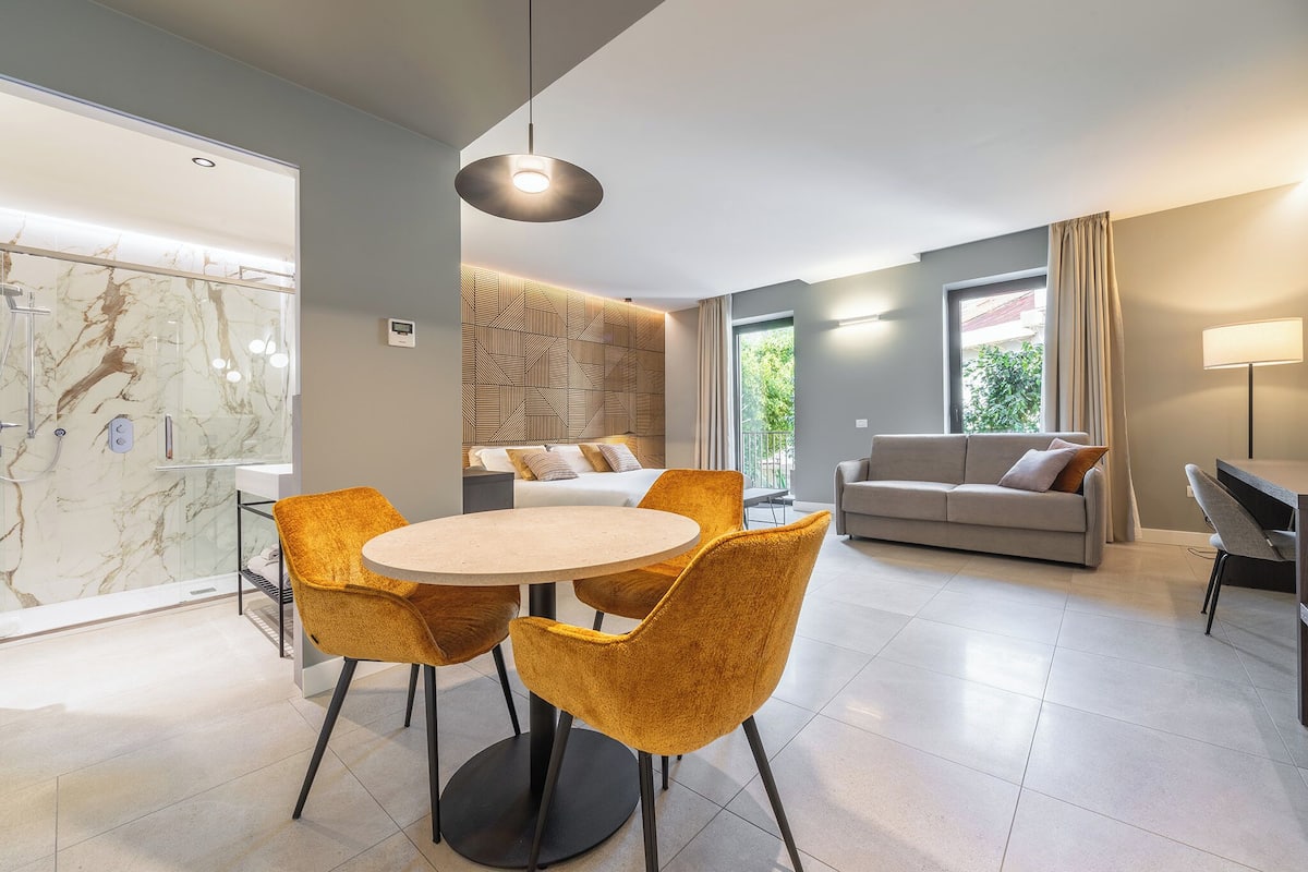 A modern living room with a round dining table, mustard-colored chairs, a sofa, and a large window with curtains.