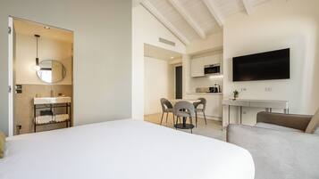 A modern hotel room with a large bed, a round mirror, a small kitchenette, a dining area with chairs, and a flat-screen TV mounted on the wall.