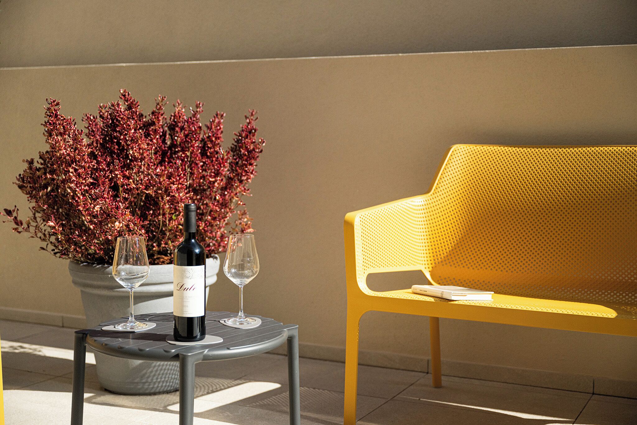A table with a bottle of wine and two glasses, a potted plant, and a yellow chair.