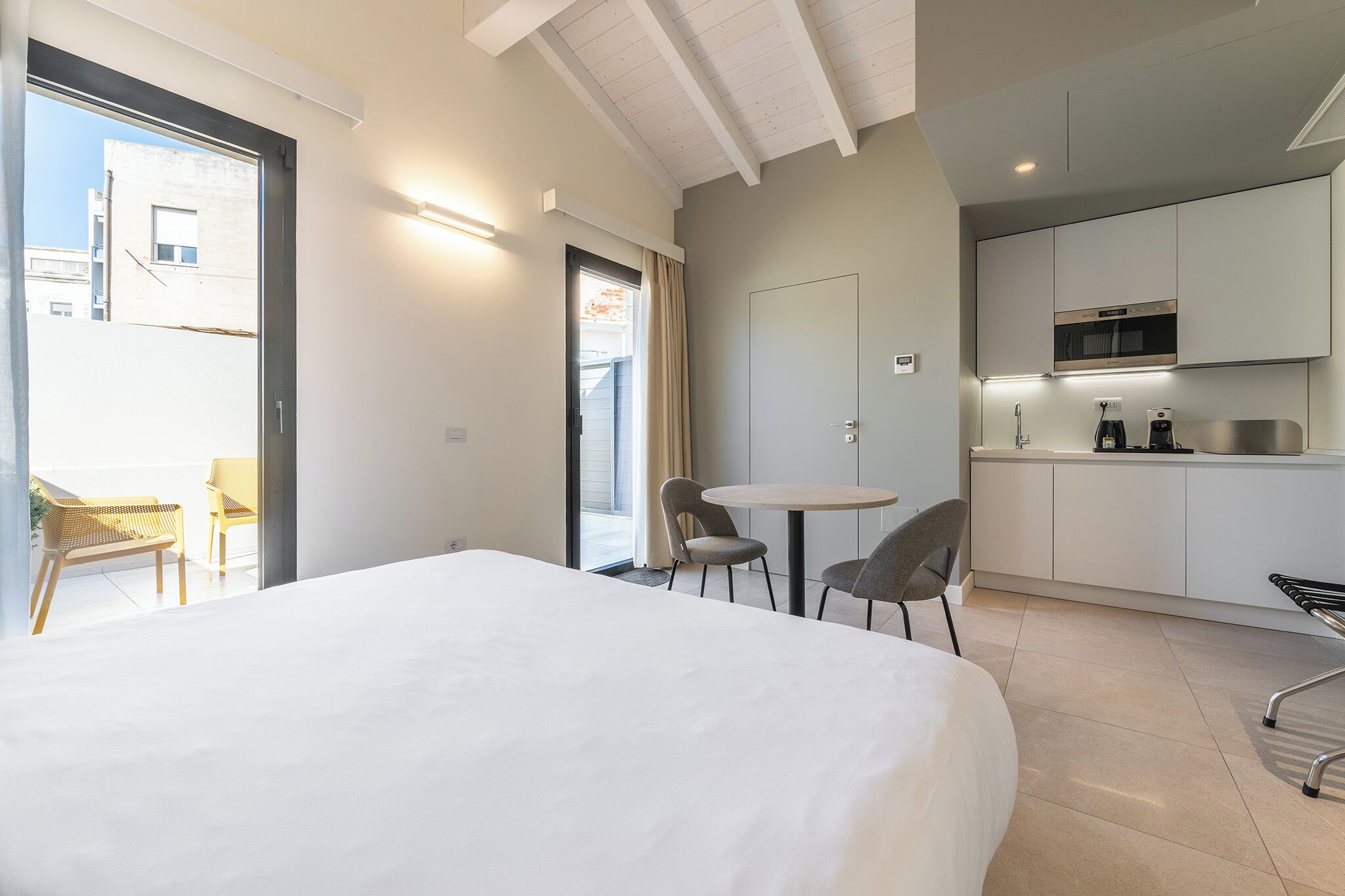 A modern hotel room with a large bed, a small kitchen area, and a balcony with seating.