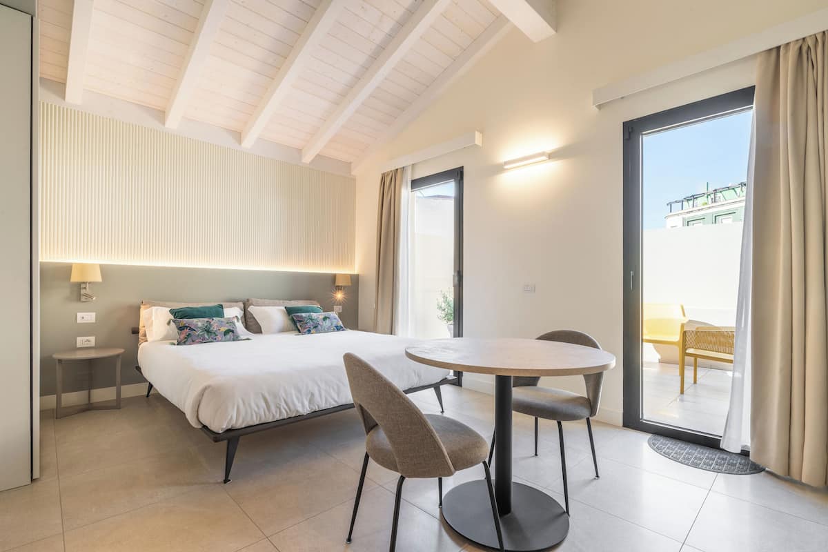 A modern hotel room with a bed, a round dining table, two chairs, a small side table, a lamp, and a view of the outside balcony.
