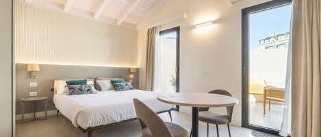 A modern hotel room with a bed, a round dining table, two chairs, a small side table, a lamp, and a view of the outside balcony.