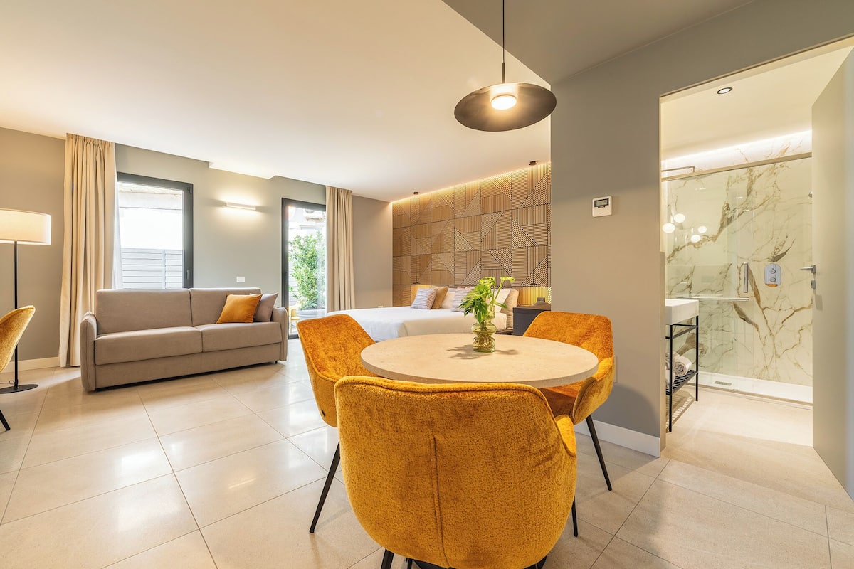 A modern hotel room with a sofa, a round dining table with chairs, a bed, and a bathroom with a glass shower.