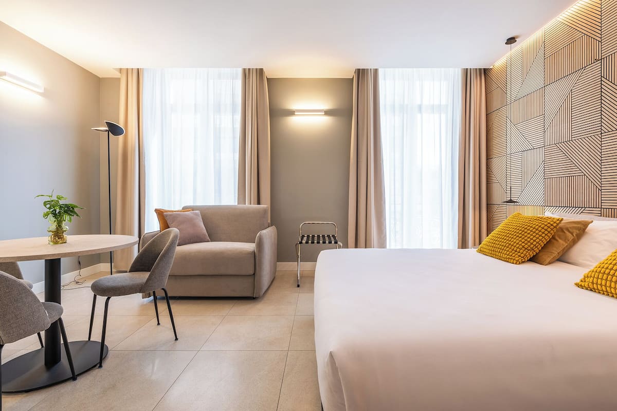 A modern hotel room with a bed, a sofa, a small table, and a chair. There is a lamp, a plant, and a window with curtains.