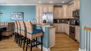 Una cocina con armarios de colores claros, un refrigerador de acero inoxidable, un microondas y una zona de comedor con sillas de mimbre y un cojín azul.