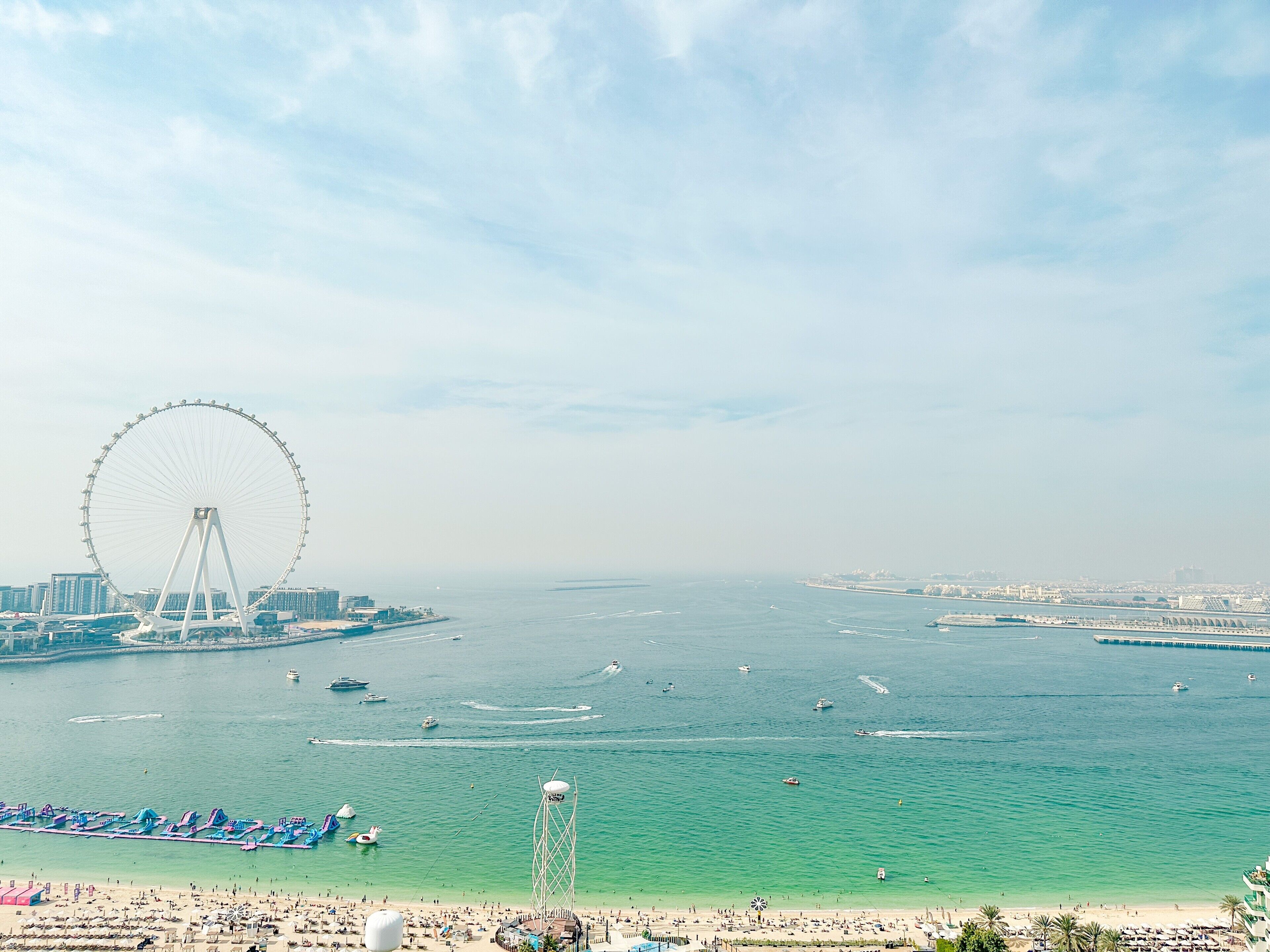 Un paysage urbain côtier avec une grande roue bien visible, une plage avec des transats et un port de plaisance rempli de bateaux.