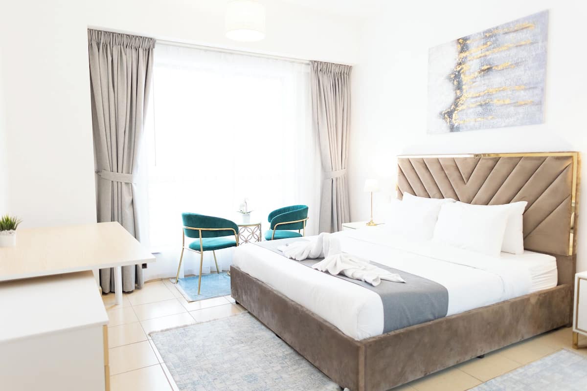 A hotel room with a large bed, a desk, two chairs, a small table, and a large window with curtains.
