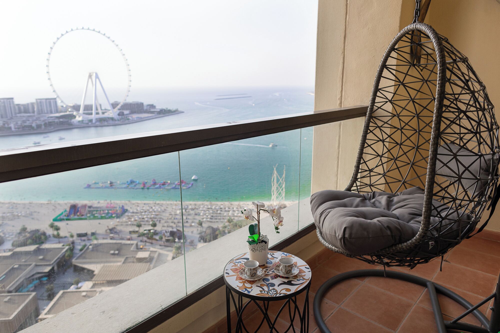 A balcony with a hanging chair, a small table with cups and a plant, and a view of a Ferris wheel and waterfront.