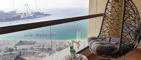 A balcony with a hanging chair, a small table with cups and a plant, and a view of a Ferris wheel and waterfront.