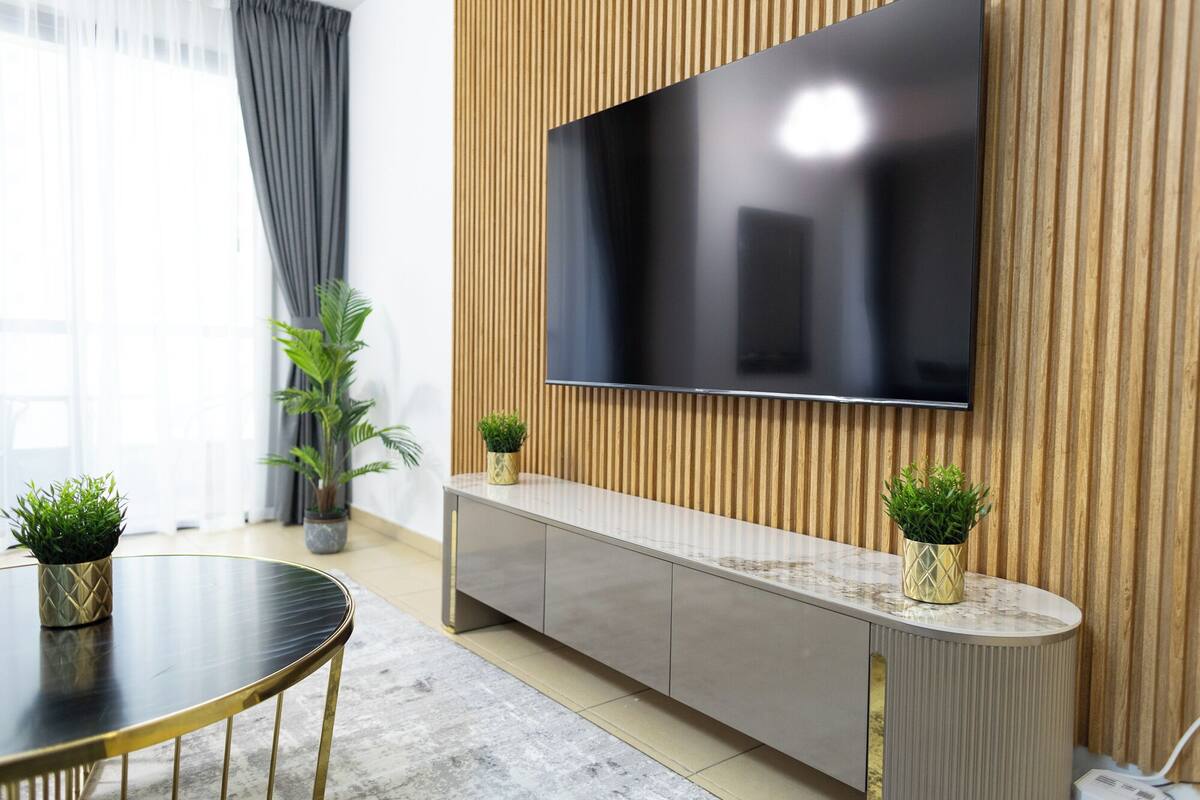 A modern living room with a large flat-screen TV mounted on a wooden wall, a grey sofa, a round dining table with gold legs, and two potted plants.