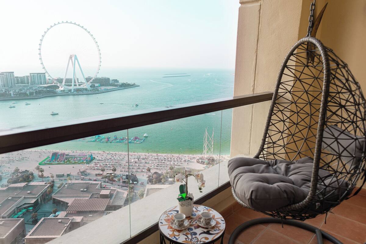 A balcony with a hanging chair, a view of a beach, and a Ferris wheel in the distance.