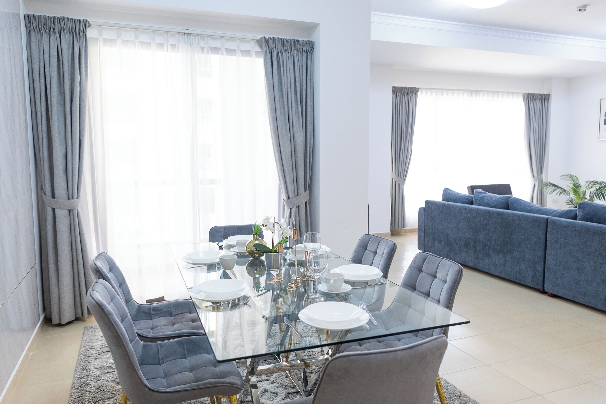 A modern dining area with a glass table and grey upholstered chairs, a blue sofa in the background, and grey curtains on the windows.
